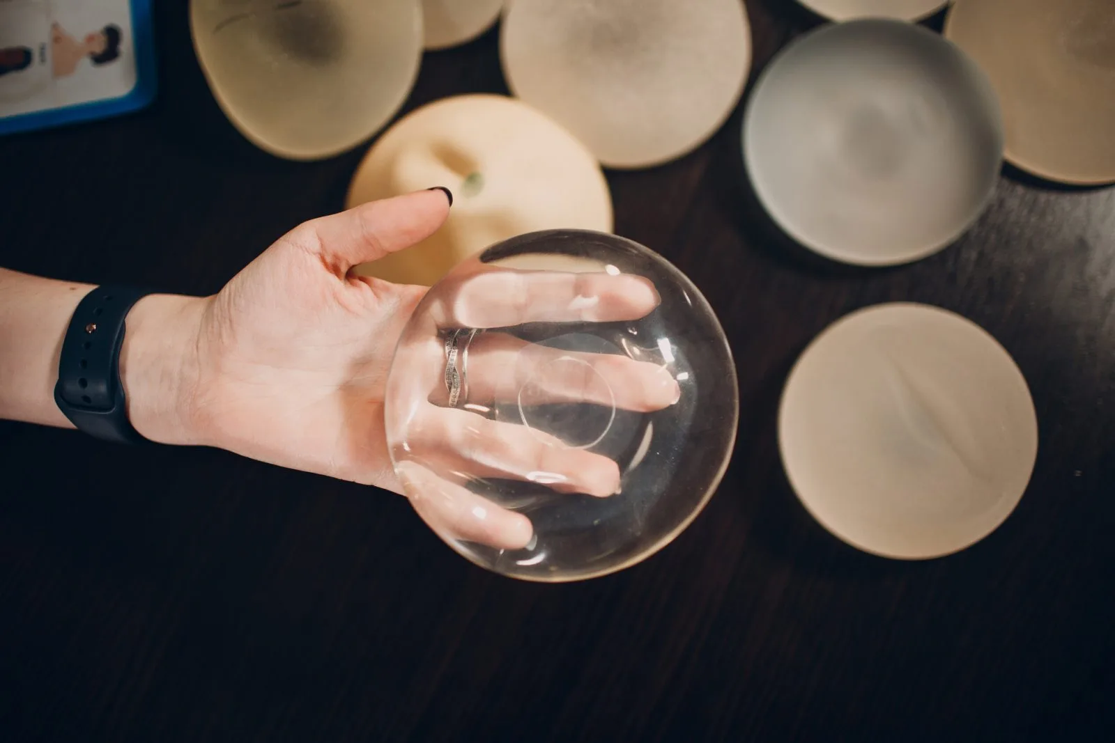 Hand holding a clear silicone breast implant with additional implants displayed on dark surface